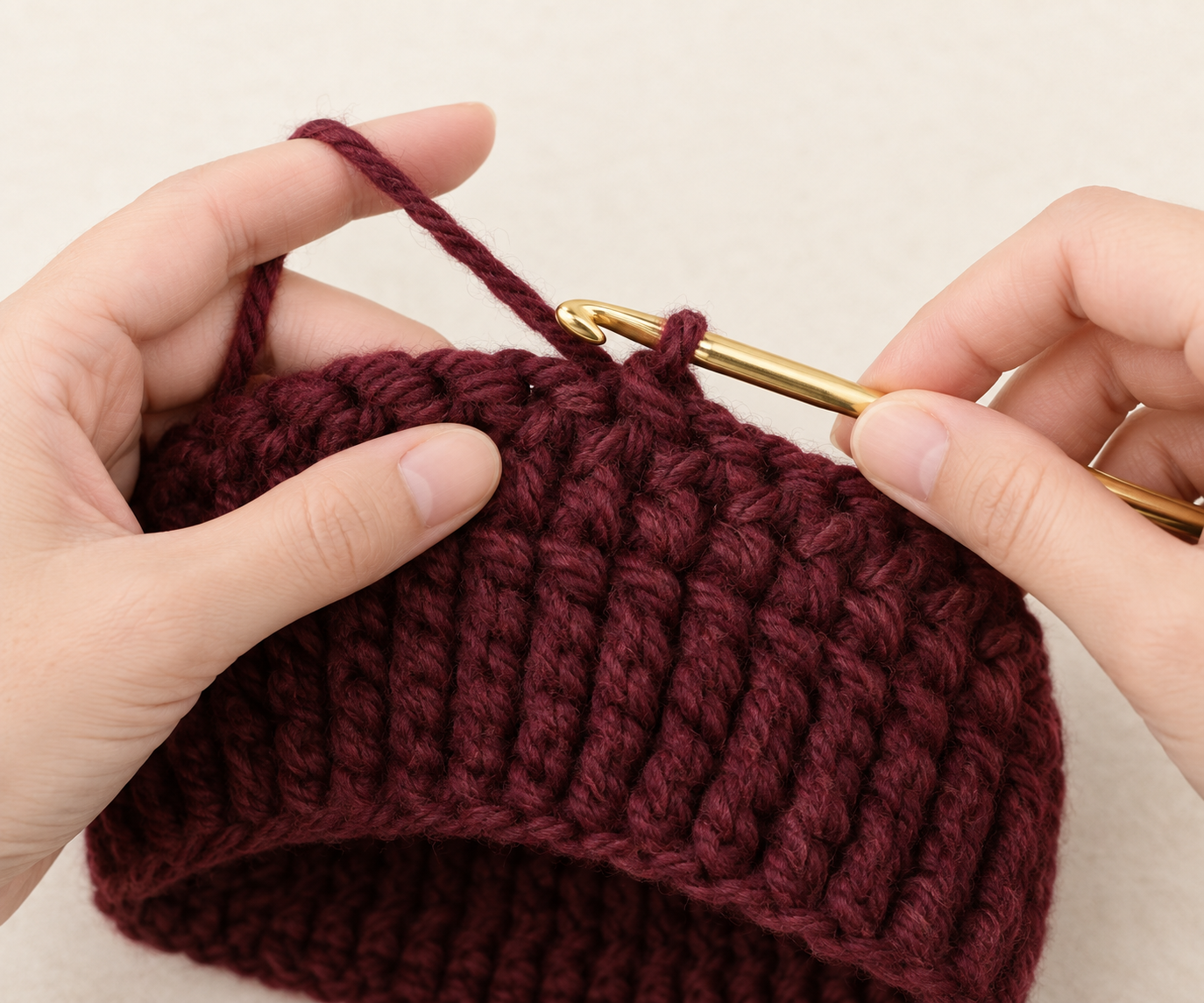 Gold crochet hook entering the top of a stitch on a burgundy textured crochet beanie body
