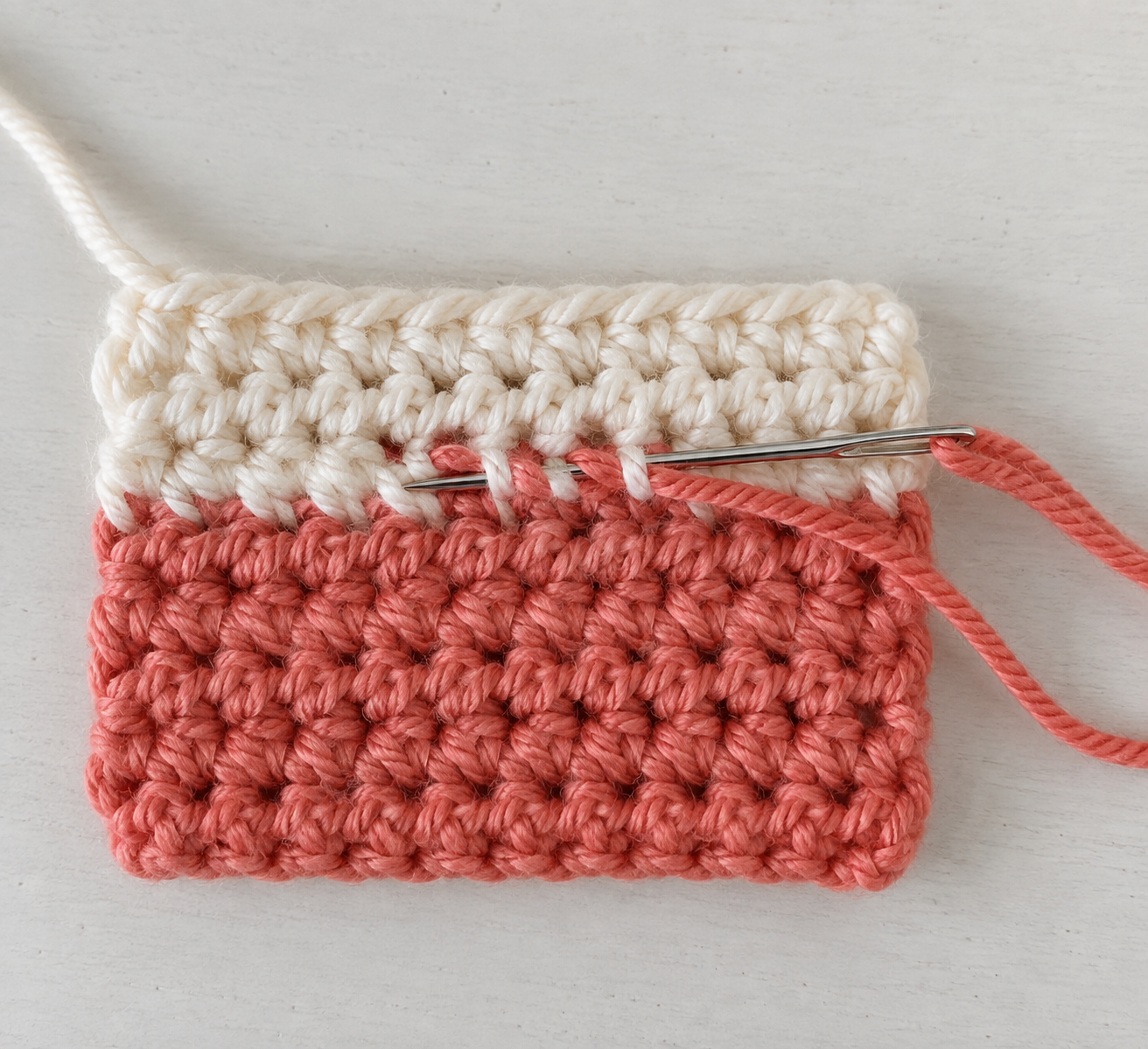 Yarn needle weaving a coral yarn tail through the wrong side of a two-color crochet swatch